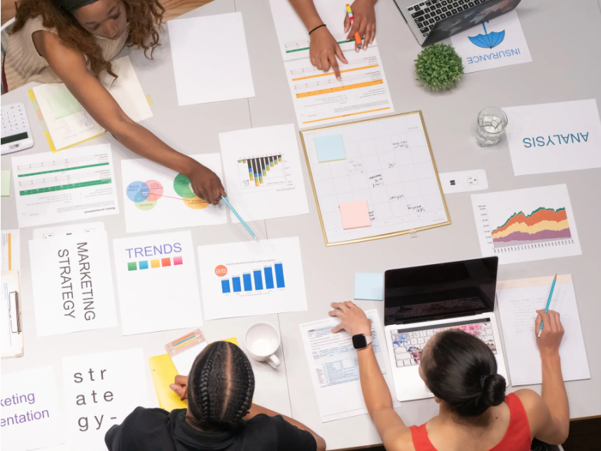 Equipo de trabajo colaborando en una mesa llena de gráficos, informes y laptops, enfocados en estrategias de marketing y análisis de datos.